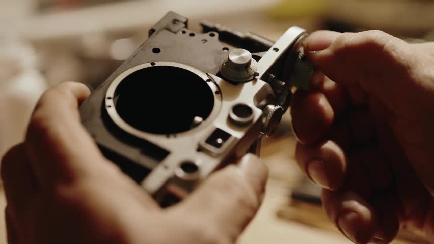 Close up of the hands of a technician inspecting a disassembled silver camera body and its internal mechanisms