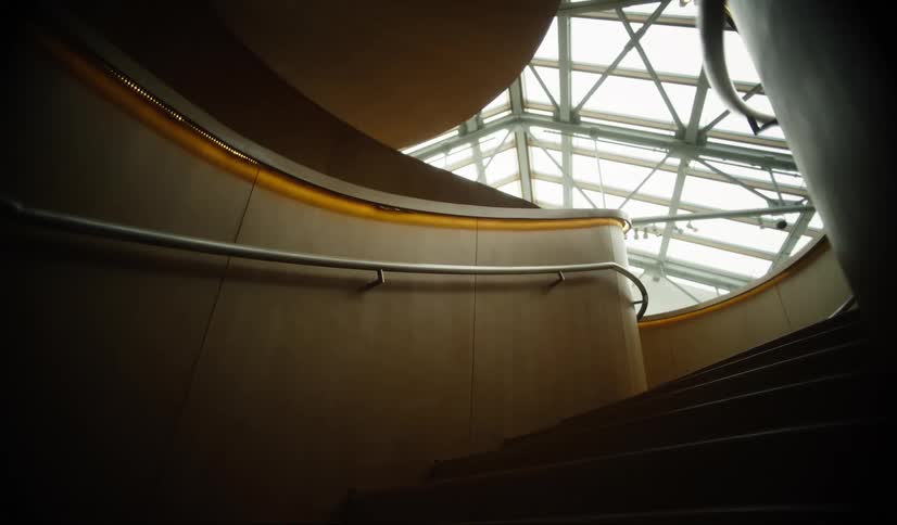 Curved wooden staircase leads to a space illuminated by a glass and metal structure on the ceiling