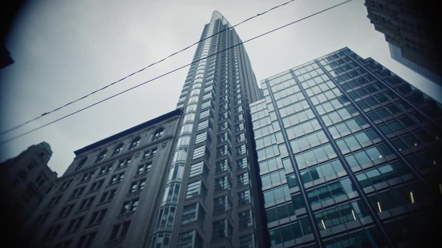 A lowangle view of several tall buildings in a city with a prominent modern skyscraper in the center flanked by other highrise structures under an overcast sky