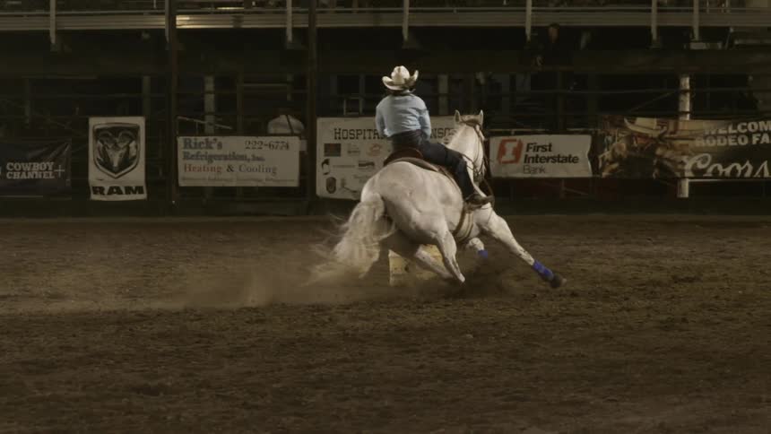 Rodeo competitor rides a white horse at high speed as it goes around a barrel in the arena