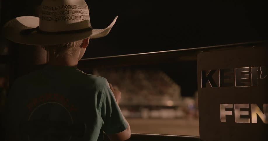 Boy in a cowboy hat watches intently at a rodeo at night