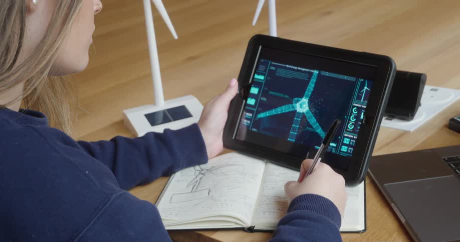 Woman examines a digital model of a wind turbine on a tablet while taking notes in her notebook