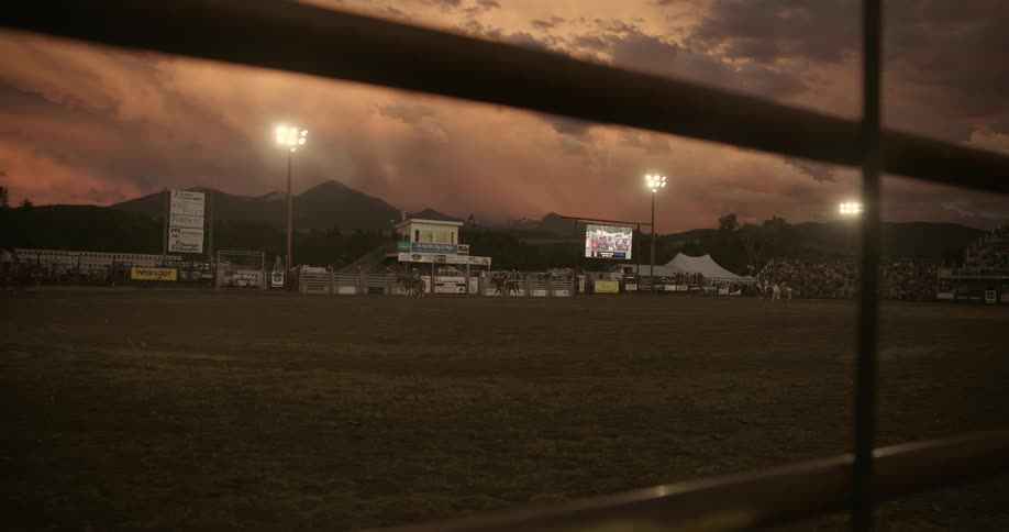 Rodeo arena at dusk seen through a fence with full stands and artificial lighting