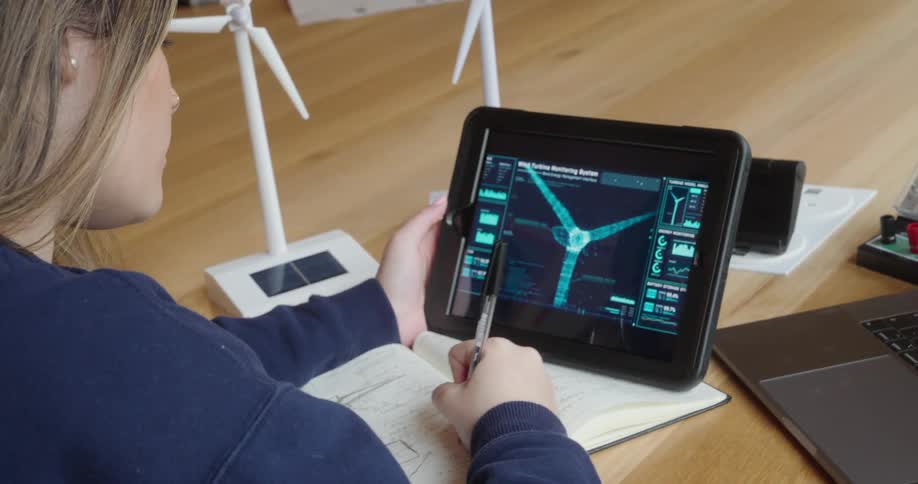 Woman analyzes data about wind turbines on a tablet while taking notes in a notebook