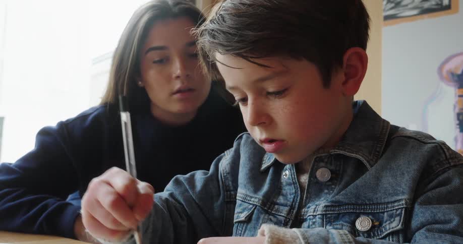 A teenager is watching a child write in a notebook