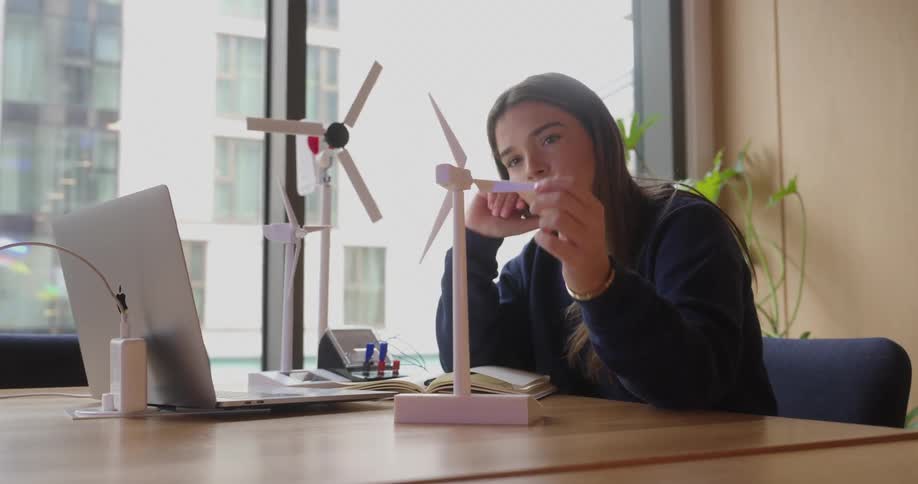 A person is sitting at a desk with a laptop two small wind turbine models and some books in a room with large windows and potted plants