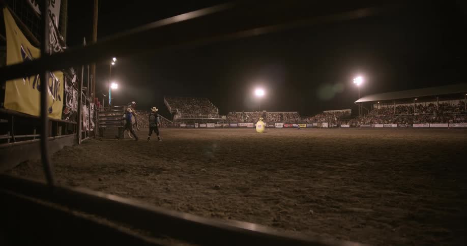 A rodeo event unfolds under bright floodlights with a bull rider gripping tightly as the bull kicks up dirt while the crowd watches from packed grandstands