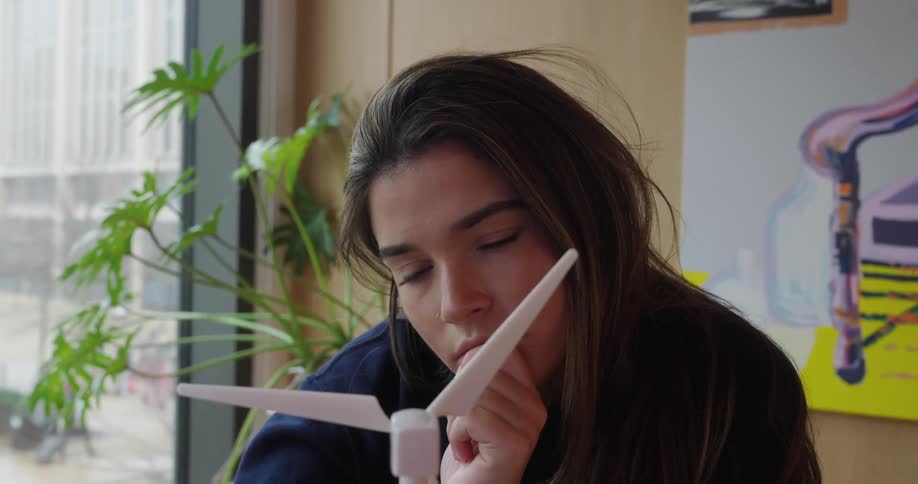 Woman with long hair looks closely at a model of a wind turbine in an indoor setting with plants and windows in the background