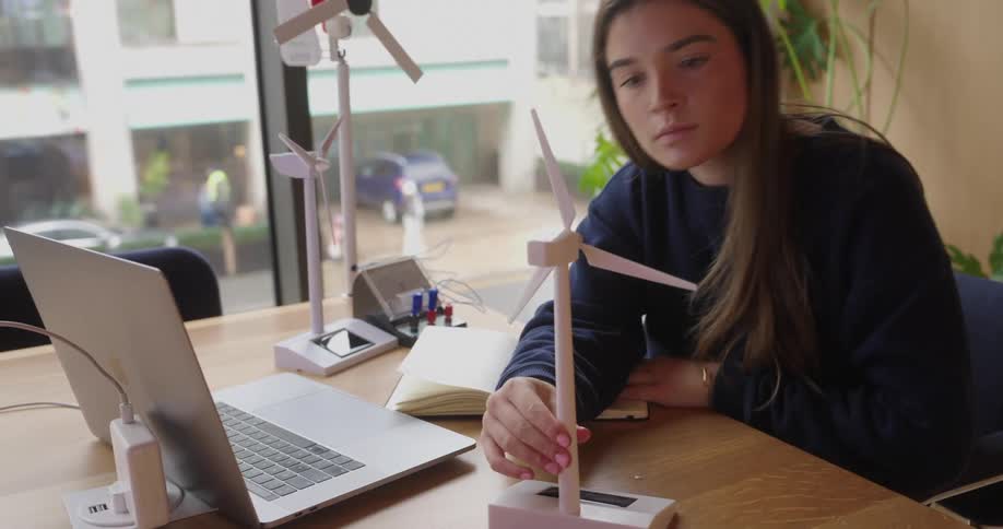 Woman handles a model wind turbine at a table with a laptop notebook and electrical equipment near a window
