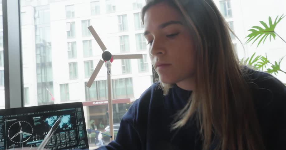 A young woman works with a miniature windmill while typing on a laptop in a conference room
