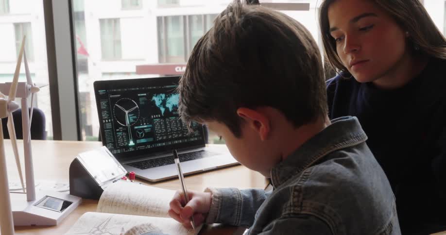 Boy writes something down in a notebook while a young woman watches intently surrounded by models of wind turbines and a laptop with technical graphics