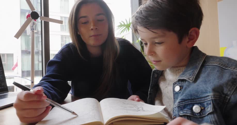 A young woman is teaching a little boy pointing the pen at a white notebook and the boy is restless