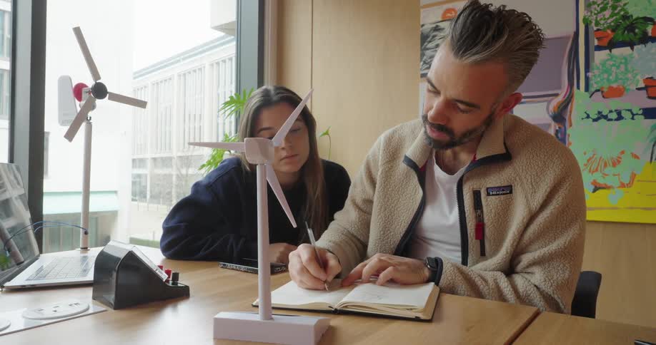 Man writes in a notebook while a young woman watches intently nearby with models of wind turbines on the table