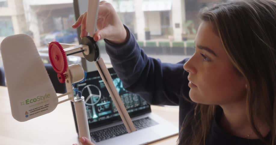 A young woman closely examines and adjusts the blades of a small model wind turbine with a laptop displaying schematics in the background