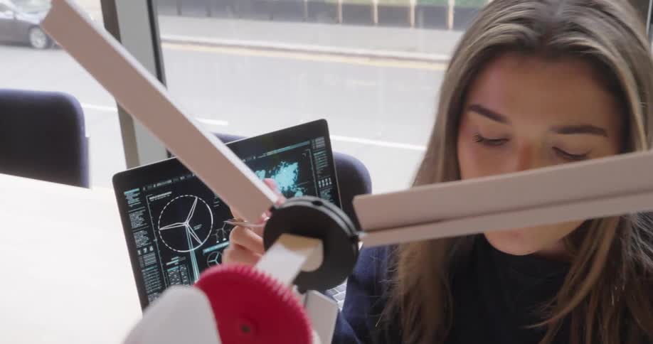 The face of a young woman who is spinning a device with three white propellers and then looking at the screen of a laptop