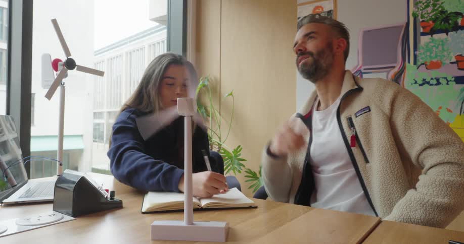 Young man writes down information in a notebook while talking to a man next to him surrounded by models of wind turbines