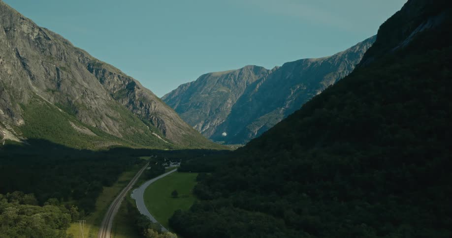 A winding road cuts through a lush valley framed by steep rocky mountains under a clear sky
