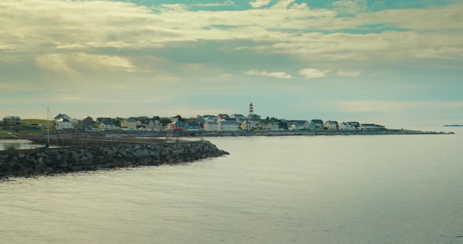 A seaside town stretches along the coastline under a partly cloudy sky viewed from a calm body of water with a rocky breakwater in the foreground