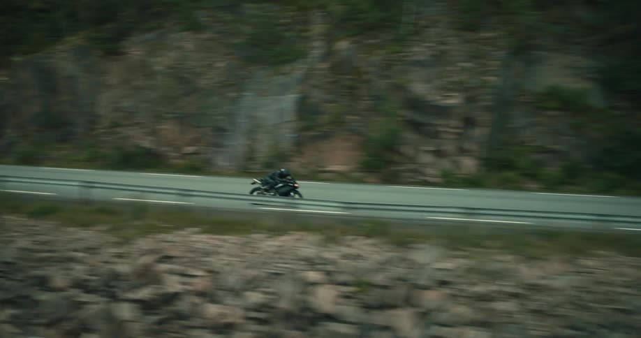 A person rides a motorcycle on a rural highway beside a river and a forest