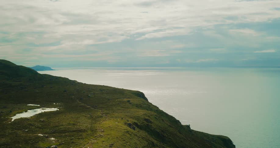 A coastal landscape with a green hillside sloping down to a calm sea under a cloudy sky
