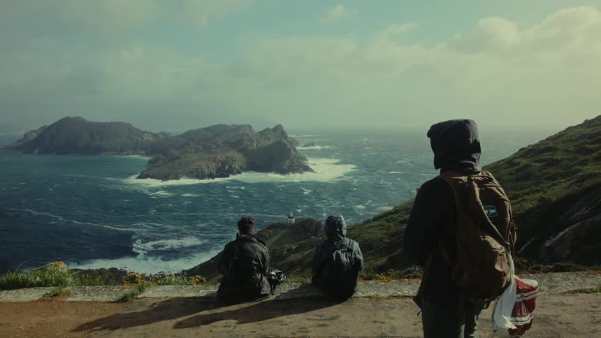 Two hikers are sitting down while another stands on a dirt road above a rocky ocean coastline