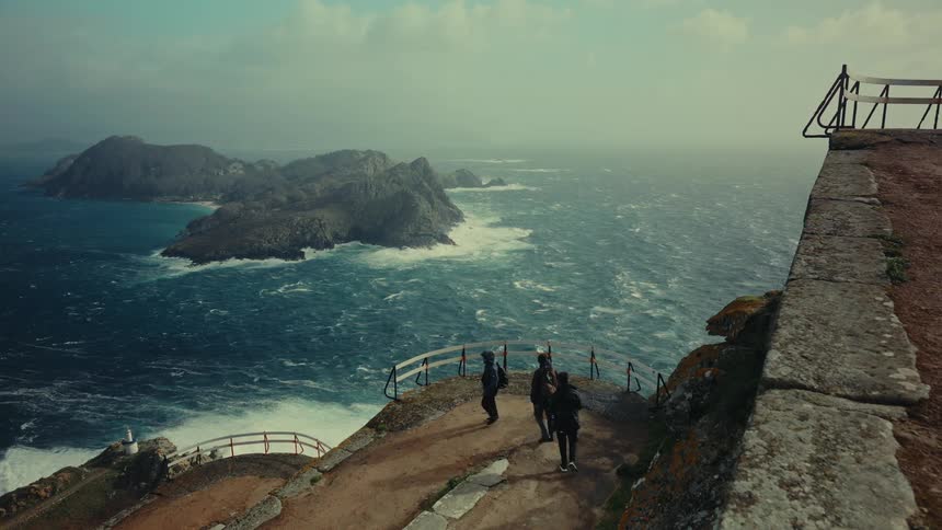 Group of people walk along a viewpoint by the ocean with rocky islands in the background and strong winds stirring the waters