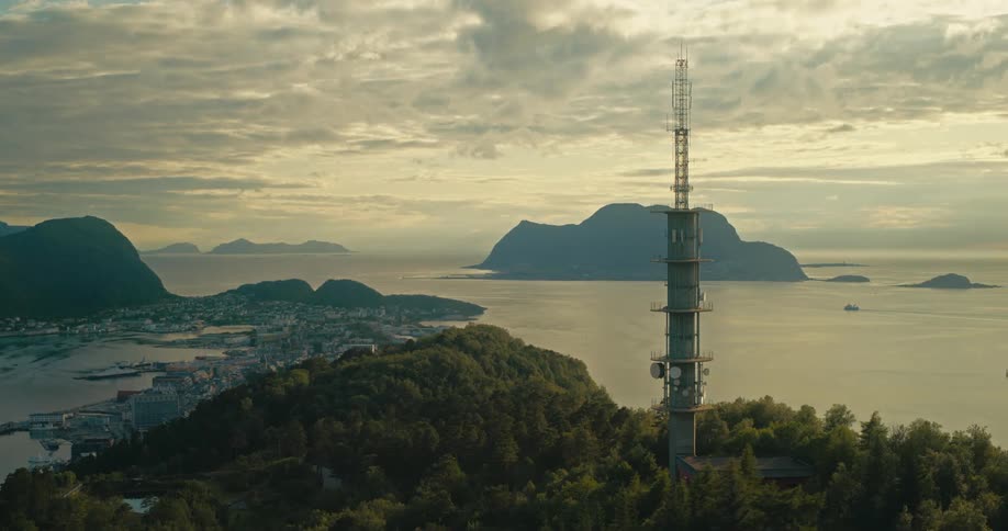An aerial view captures a tall tower overlooking a cityscape nestled by water with mountains and islands visible in the distance under a partly cloudy sky