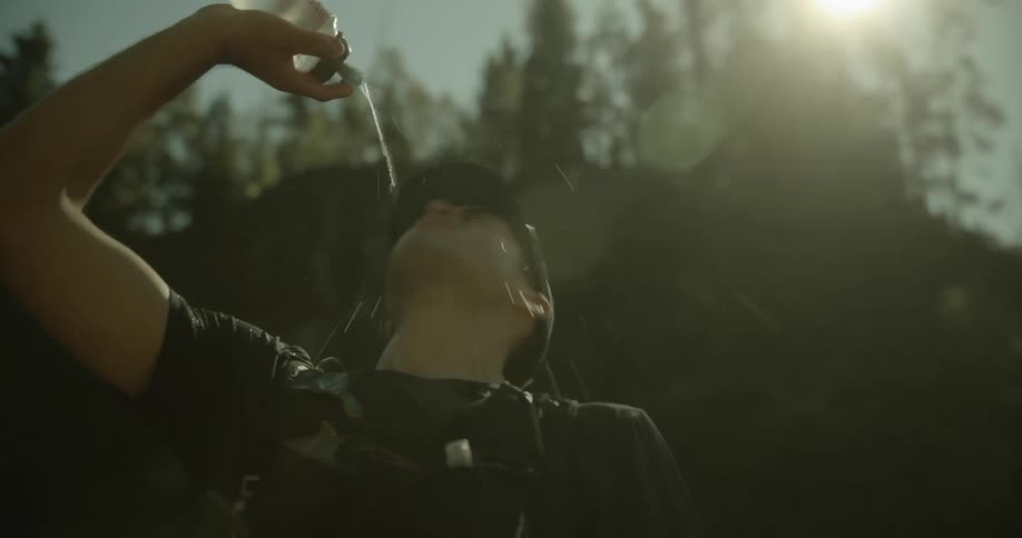 Man standing near the base of a forested hill on a sunny day pours water onto his face from a water bottle