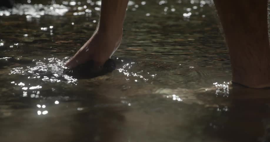 A man is washing their hat in a flowing river dipping and shaking it in the water before picking it back up with sunlight reflecting on the rivers surface showcasing a moment of riverside cleaning