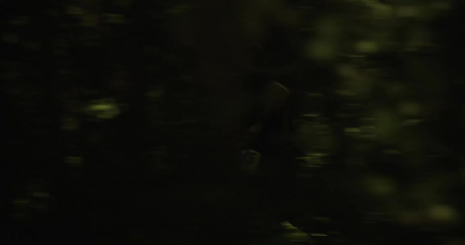A man is energetically running through dense woods in a race indicated by a tracking number highlighting the challenging shadowy and foliagerich woodland environment