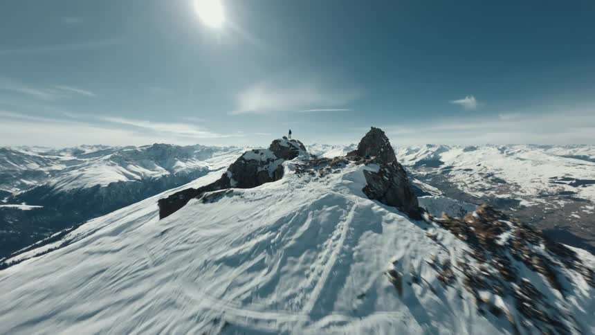 A man stands on a snowy mountain peak under a bright sun surrounded by a landscape of rugged snow covered ridges and towering rock formations