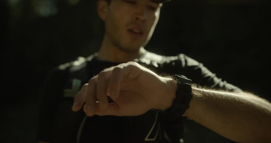 A man wearing a cap goggles and a backpack looks off camera then checks his watch in a dimly lit outdoor setting
