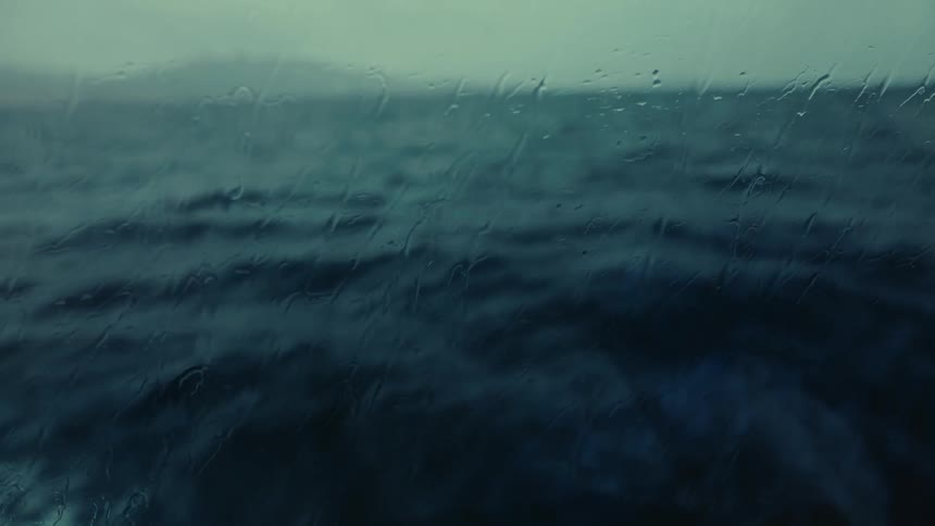 View of the ocean under a cloudy sky is captured through a glass with raindrops