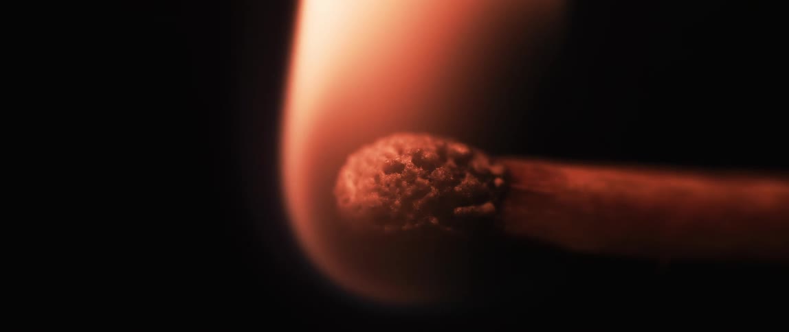A closeup image of a matchstick head igniting with the flame illuminating the textured surface of the match head against a dark background