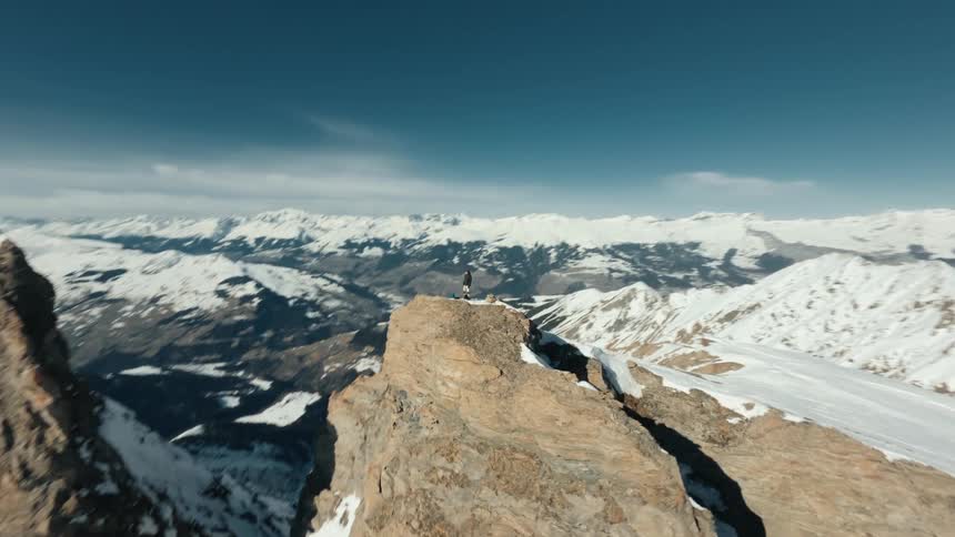A lone hiker stands on a snowy mountain peak surrounded by a vast expanse of rugged snowcovered mountains under a clear blue sky