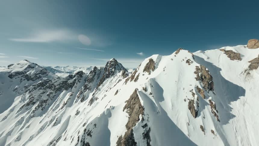 An aerial view of snow covered mountain peaks under a bright blue sky with a few wispy clouds and a lens flare visible