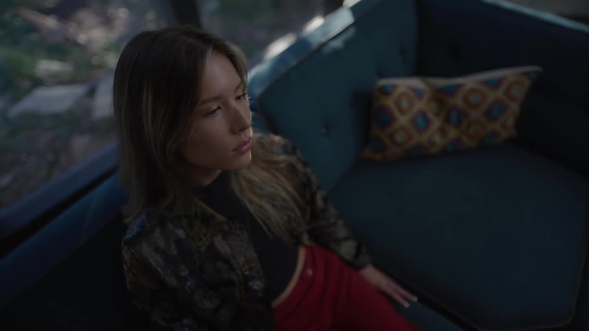 A young woman with long brown hair sits on a dark blue couch gazing thoughtfully into the distance