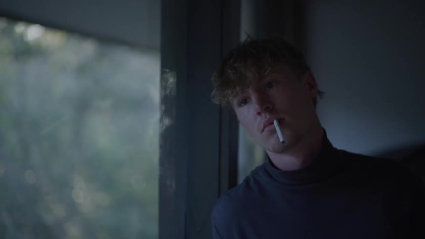 Man leans his head against the window with a cigarette in his mouth and a distant expression