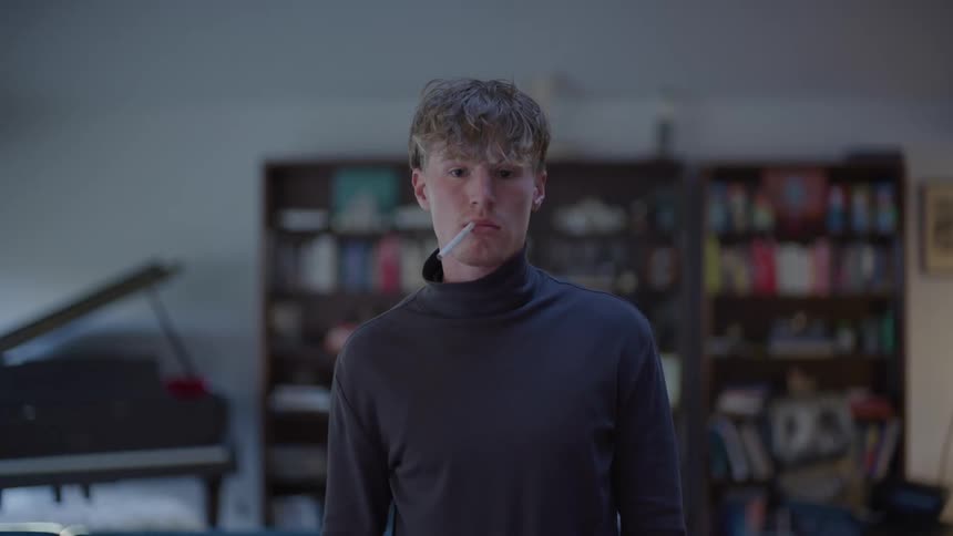 Man in dark sweater and neutral expression holds a cigarette in his mouth in a room with bookshelves and a piano in the background