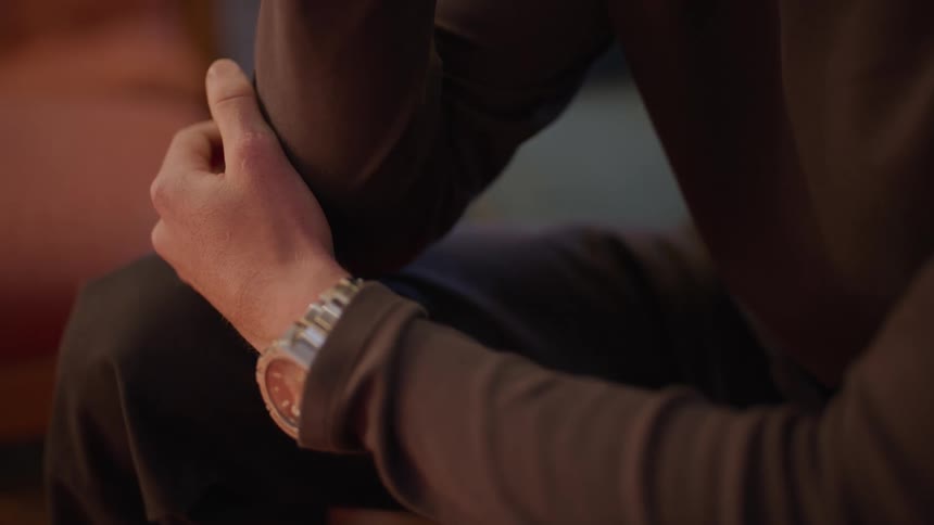 A mans hand resting on their knee wearing a stylish silver wristwatch with a soft and warm lighting ambiance