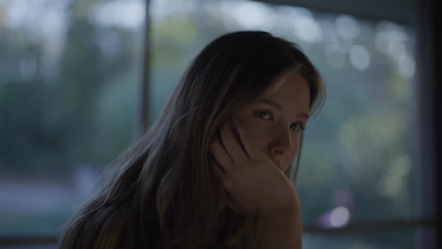 Woman rests her face on her hand and looks thoughtfully ahead with a blurred window in the background