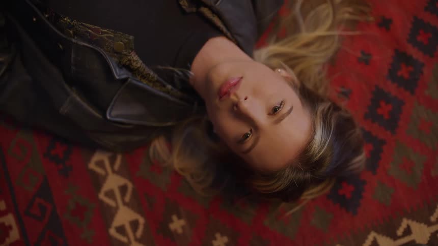 A woman lies on a bright red patterned carpet gazing upward in a relaxed thoughtful pose with her blonde hair spread out around her