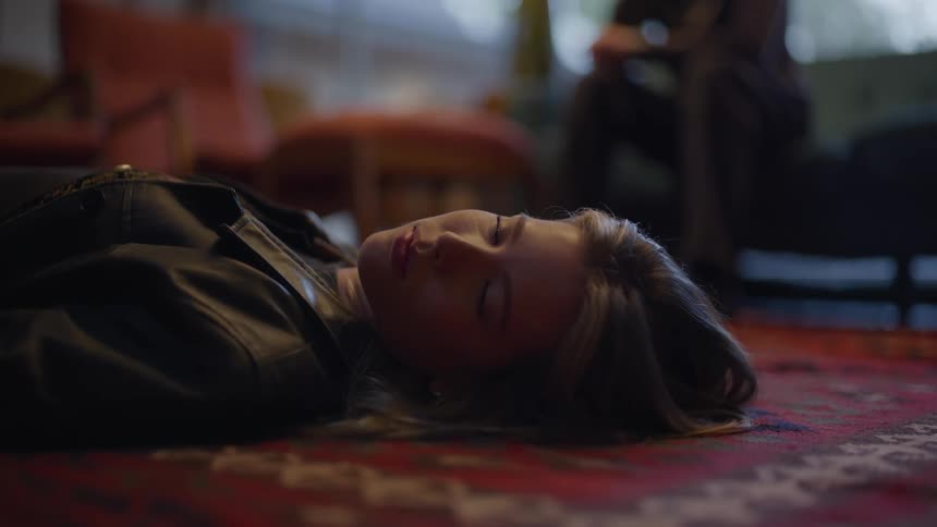 A woman looks over as she lays on a floor and someone watches from a couch