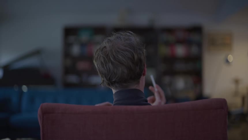 A man with slightly messy hair sits in a red chair holding a cigarette while facing away from the camera with a dimly lit room and a blurred bookshelf in the background