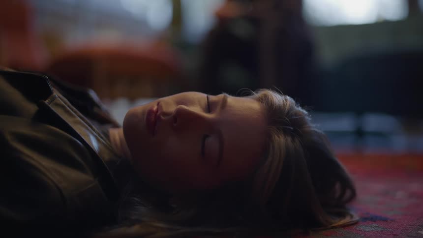 A woman with blonde hair lies on the floor with her eyes closed wearing a black leather jacket while the blurred background suggests an indoor setting