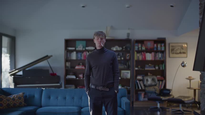 A young man in longsleeved clothing standing looking forward with a cigarette in his mouth in a living room
