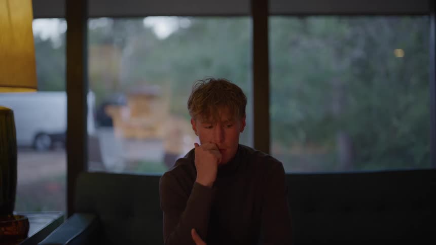 A young man looking worried is thinking while sitting on a sofa near a large window