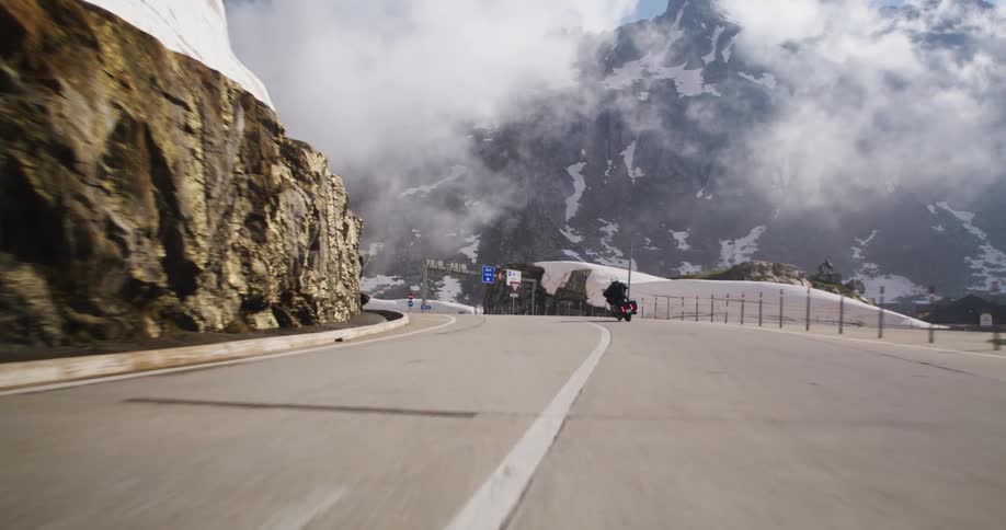 Motorcyclist makes a sharp turn on a mountain road with snow and partly cloudy skies