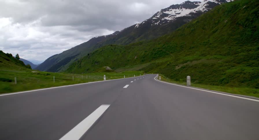 From a low streetlevel view the video pans up to follow a motorcyclist driving down a rural mountain road framed by grassy hills and distant snowcapped mountain peaks