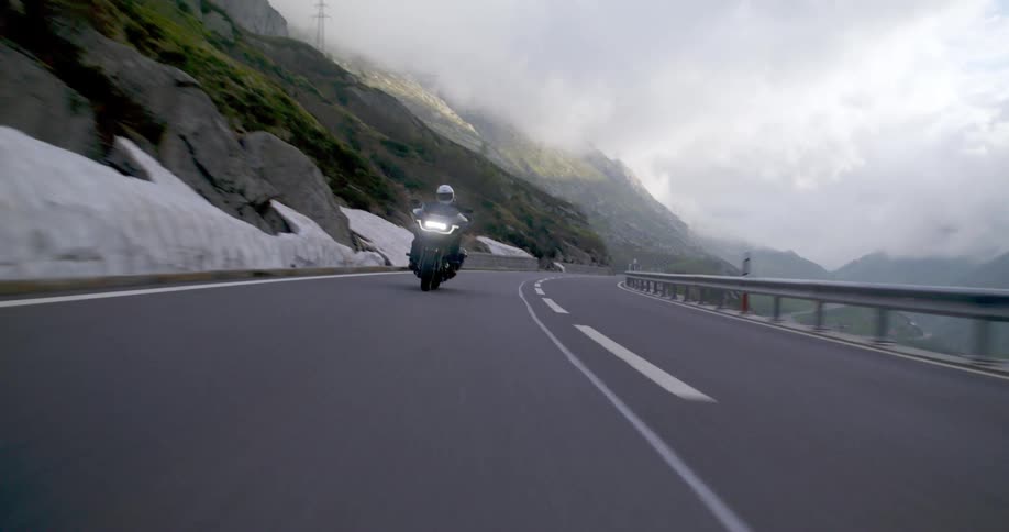 A person driving a motorcycle with the headlights on on a paved road next to a mountain